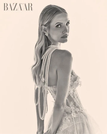 Leonie Hanne in a lace dress with layered pearl necklaces, looking back over her shoulder in a monochrome editorial portrait.