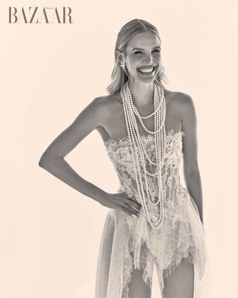 Blonde woman in strapless lace dress wearing multiple pearl necklaces laughing against a plain light background
