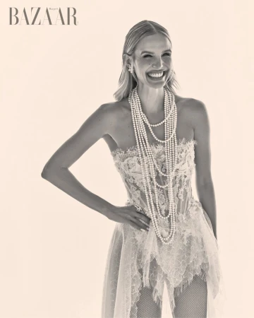 Blonde woman in strapless lace dress wearing multiple pearl necklaces laughing against a plain light background