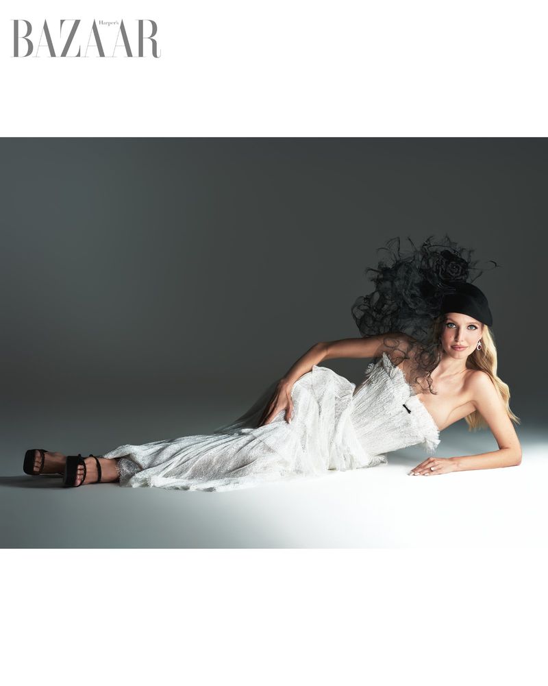Leonie Hanne lying on a grey studio floor wearing a white textured dress and black feathered fascinator for Harper's Bazaar Vietnam