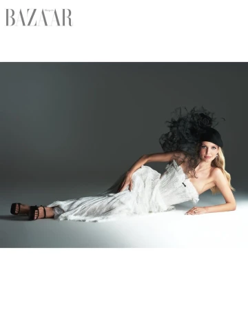 Leonie Hanne lying on a grey studio floor wearing a white textured dress and black feathered fascinator for Harper's Bazaar Vietnam