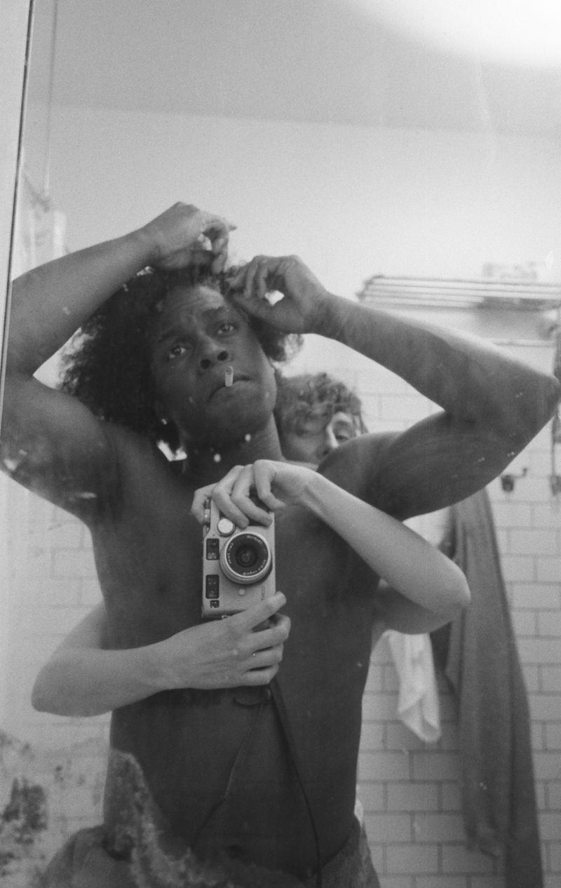Black and white mirror selfie of Daniel Caesar taking a photo with a vintage camera in a tiled bathroom.