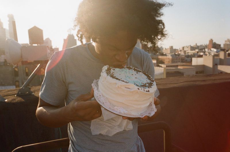 A man with dark curly hair wearing a striped t-shirt eating a frosted white cake on a rooftop at sunset.