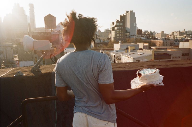 Man holding a frosted cake on a rooftop overlooking a city skyline at sunset for Interlope Magazine editorial
