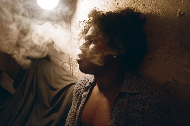 Close-up portrait of a man exhaling thick smoke indoors, wearing a textured grey knit shirt against a rough wall.