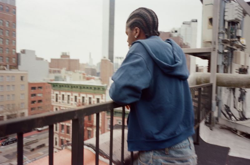 Man wearing a blue hooded sweatshirt leaning on a metal rooftop railing looking out over a city skyline