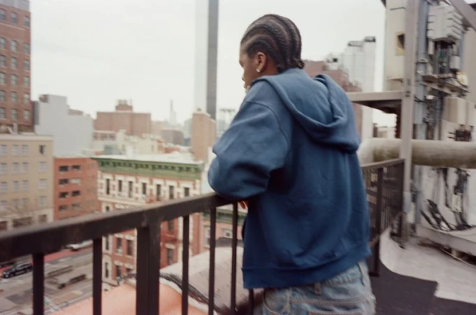 Man wearing a blue hooded sweatshirt leaning on a metal rooftop railing looking out over a city skyline
