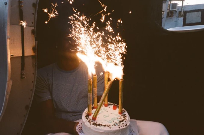 Man seated behind a birthday cake with lit sparklers for Interlope Magazine, photographed by Elena Mottola.