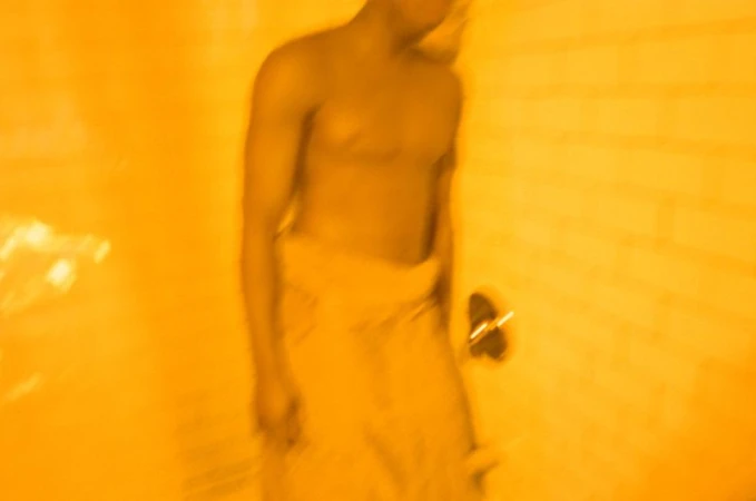 Shirtless man wrapped in a towel standing in a tiled bathroom with warm amber lighting