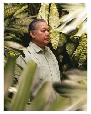 Portrait of a man wearing a light green button-down shirt framed by lush green tropical foliage and ferns.