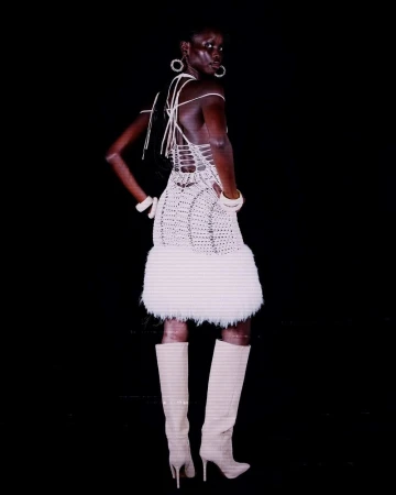 Model in white halter dress with feathered hem and knee-high boots against a black seamless background
