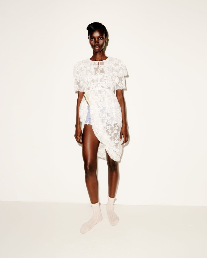 Black female model wearing a white lace dress and socks against a plain light wall background.