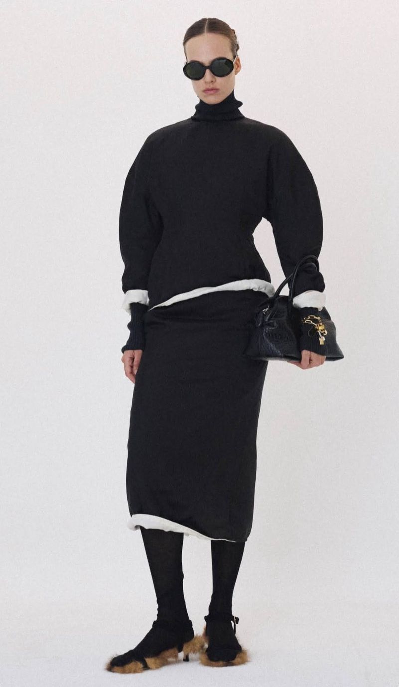 Model in dark sunglasses wearing a black turtleneck and black skirt holding a handbag against a plain white background