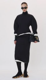 Model in dark sunglasses wearing a black turtleneck and black skirt holding a handbag against a plain white background