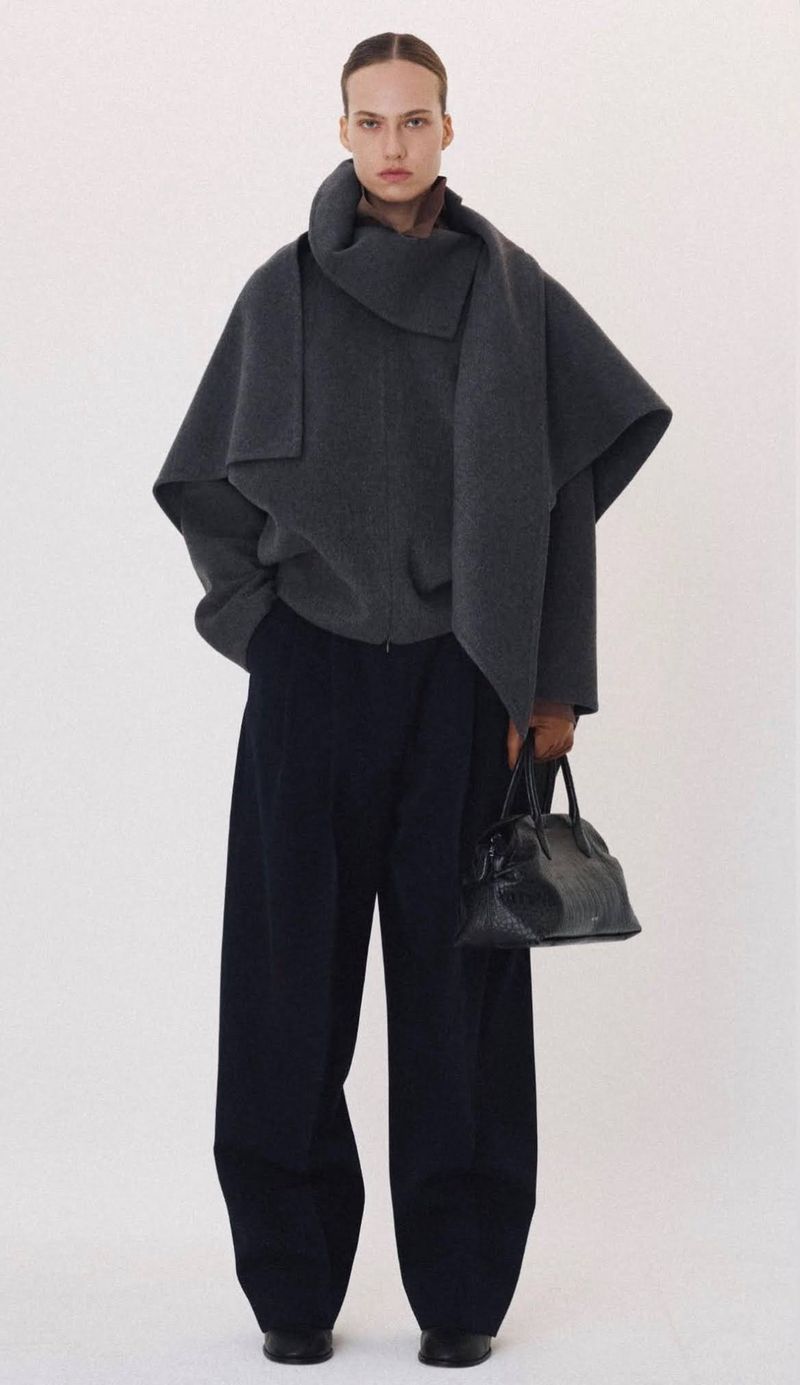Model in a dark grey wrap coat and wide trousers holding a black handbag against a plain white background
