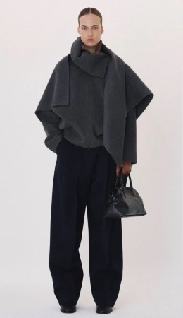 Model in a dark grey wrap coat and wide trousers holding a black handbag against a plain white background
