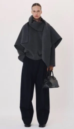 Model in a dark grey wrap coat and wide trousers holding a black handbag against a plain white background