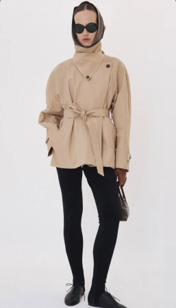 Model in dark sunglasses and headscarf wearing a tan belted coat against a white background