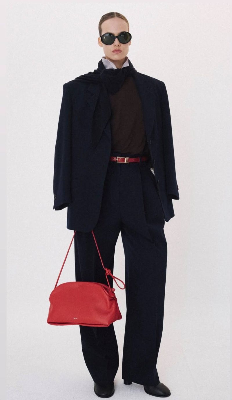 Model in dark sunglasses and dark oversized suit holding a red handbag against a bright white background for Recto Fall/Winter 2026.