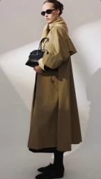 Model in dark sunglasses wearing a long beige trench coat holding a black handbag against a light background