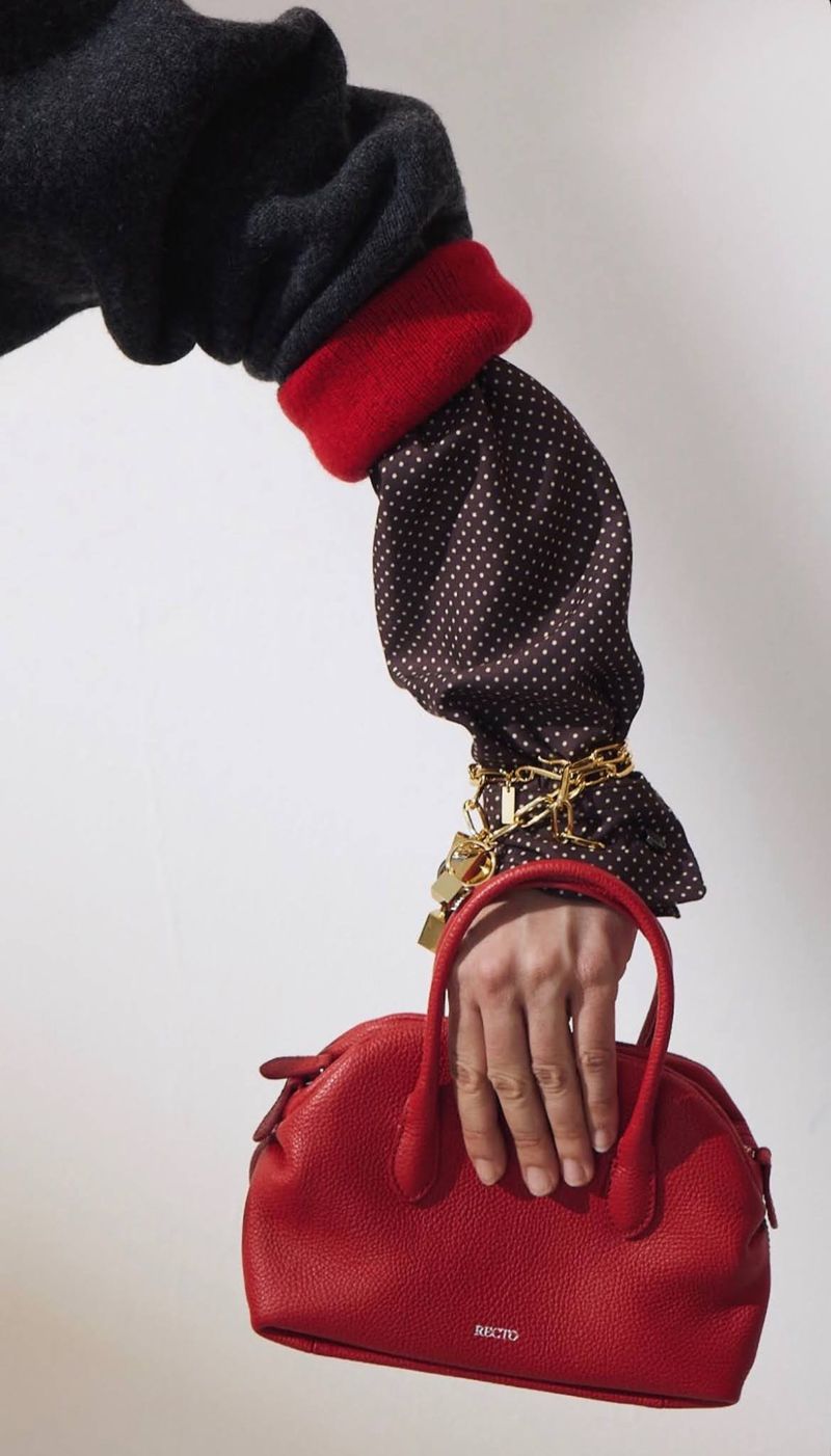Close-up of a model holding a red leather handbag, wearing a dark sweater, red cuff, polka dot sleeve, and gold chains.