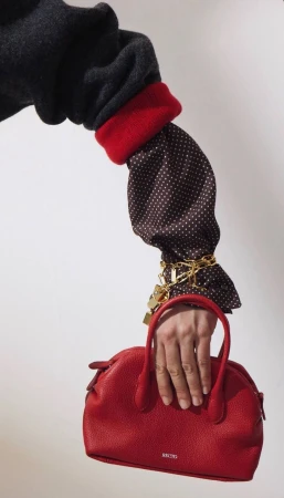 Close-up of a model holding a red leather handbag, wearing a dark sweater, red cuff, polka dot sleeve, and gold chains.