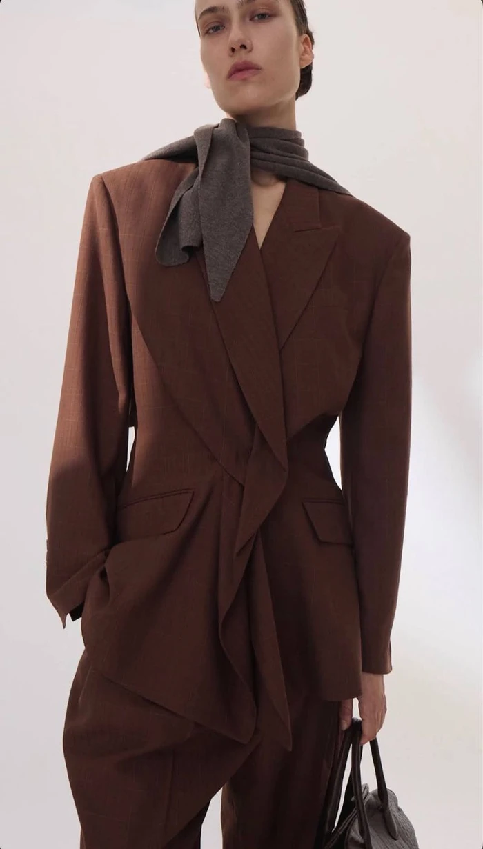 Model wearing a structured brown blazer with a grey scarf and holding a handbag against a seamless white studio backdrop.