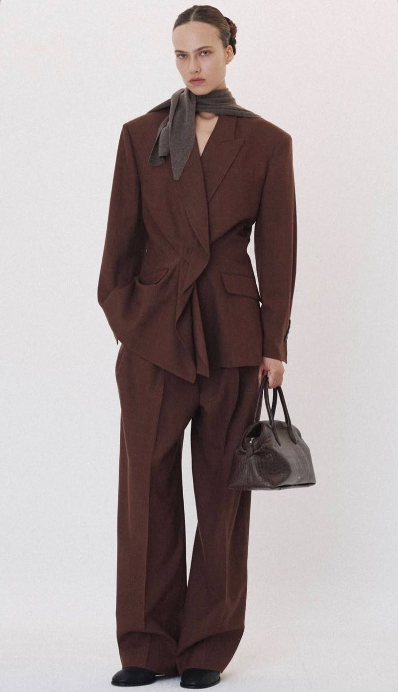 Model in brown oversized blazer and trousers with grey scarf holding dark handbag against white backdrop