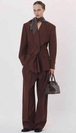 Model in brown oversized blazer and trousers with grey scarf holding dark handbag against white backdrop