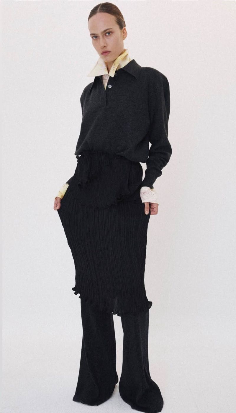 Model wearing a dark long-sleeve collared top and pleated skirt against a plain white studio background.