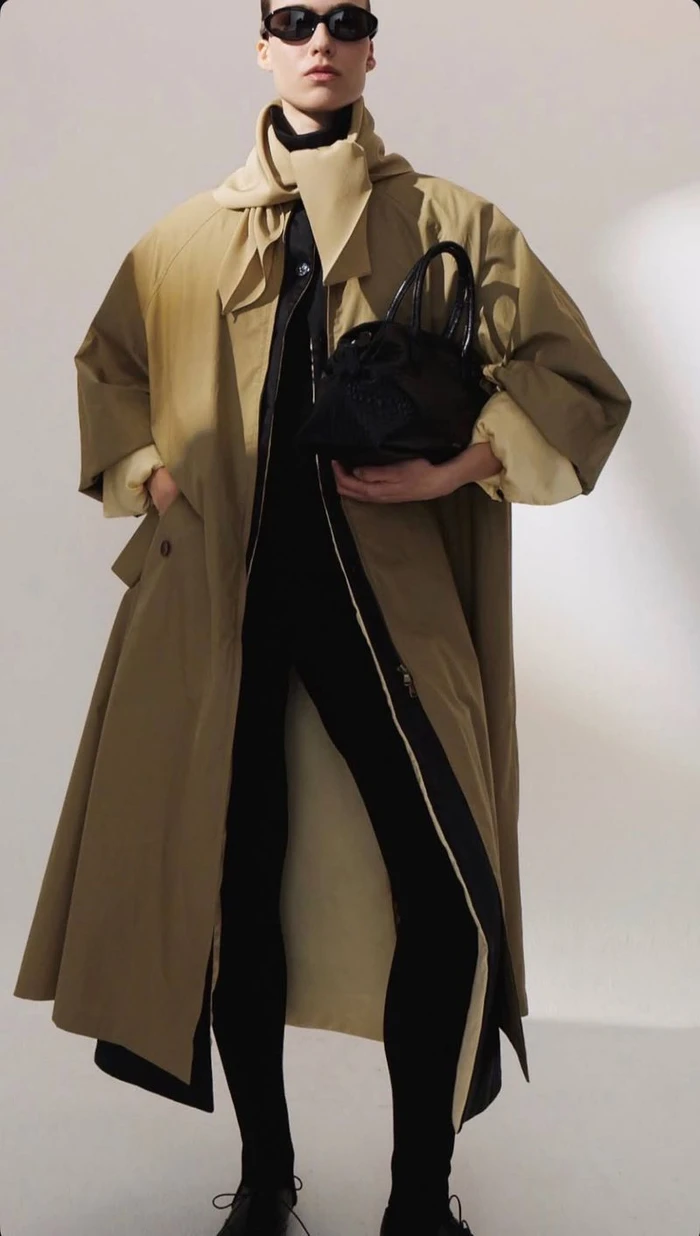 Model in dark sunglasses wearing an oversized tan trench coat and carrying a black handbag against a light studio background