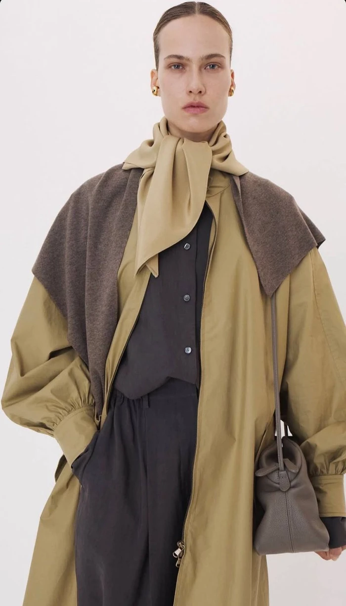 Model wearing a tan coat, layered scarf, and dark shirt against a seamless white background, holding a handbag.