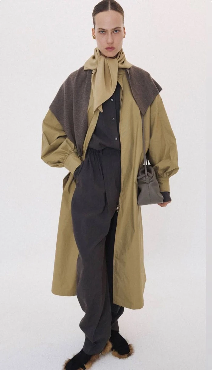 Model wearing a tan coat, dark trousers, scarf, and holding a small handbag against a plain white background.