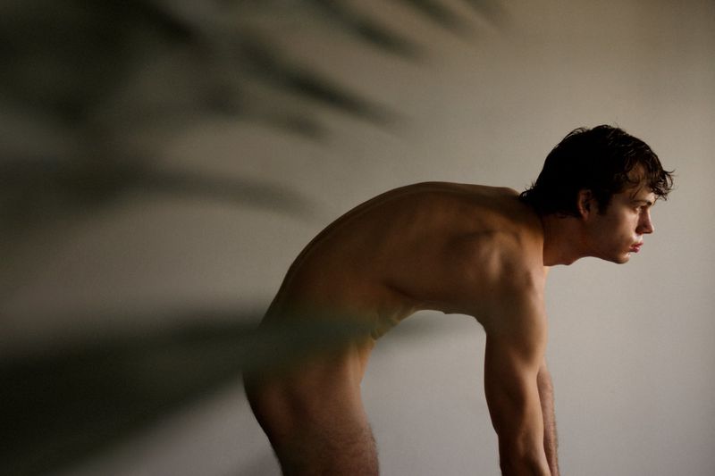 Nude man with dark hair is bent over, with his head facing right, in front of a light grey wall.  A shadow is blurred in the frame.
