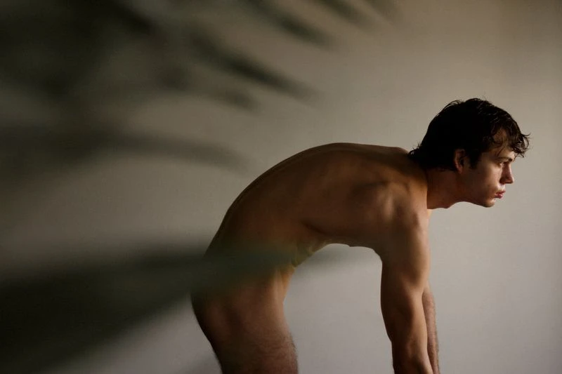 Nude man with dark hair is bent over, with his head facing right, in front of a light grey wall.  A shadow is blurred in the frame.