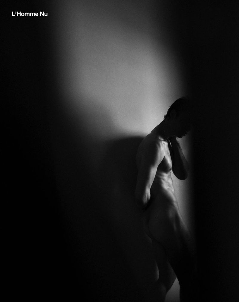 Nude male figure leaning against a wall in a black and white studio shot. The subject is partially obscured by shadow.