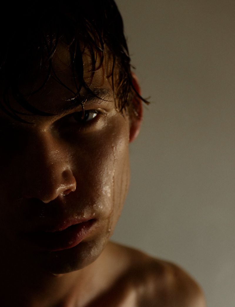 Close-up portrait of a young adult male with wet hair and skin, gazing intensely at the camera against a blurred neutral background.