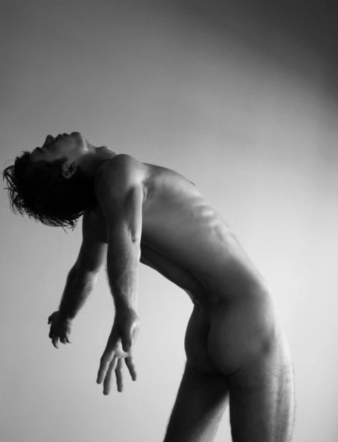 Black and white portrait of a shirtless man arching backwards with head tilted and arms dangling in front of a pale background.