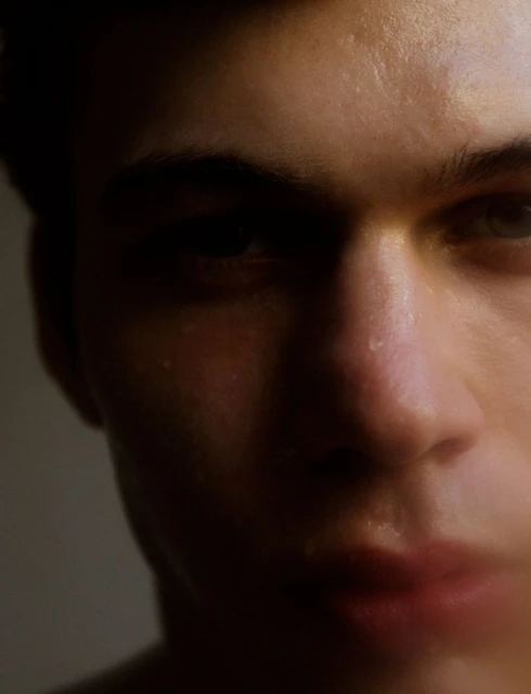 Close-up portrait of a young adult male with light skin, showing sweat droplets on his face. The lighting is dramatic and moody.