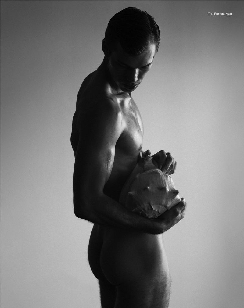 Monochrome portrait of nude adult male holding seashell in studio setting with gradient background. Low key lighting creates soft shadows.