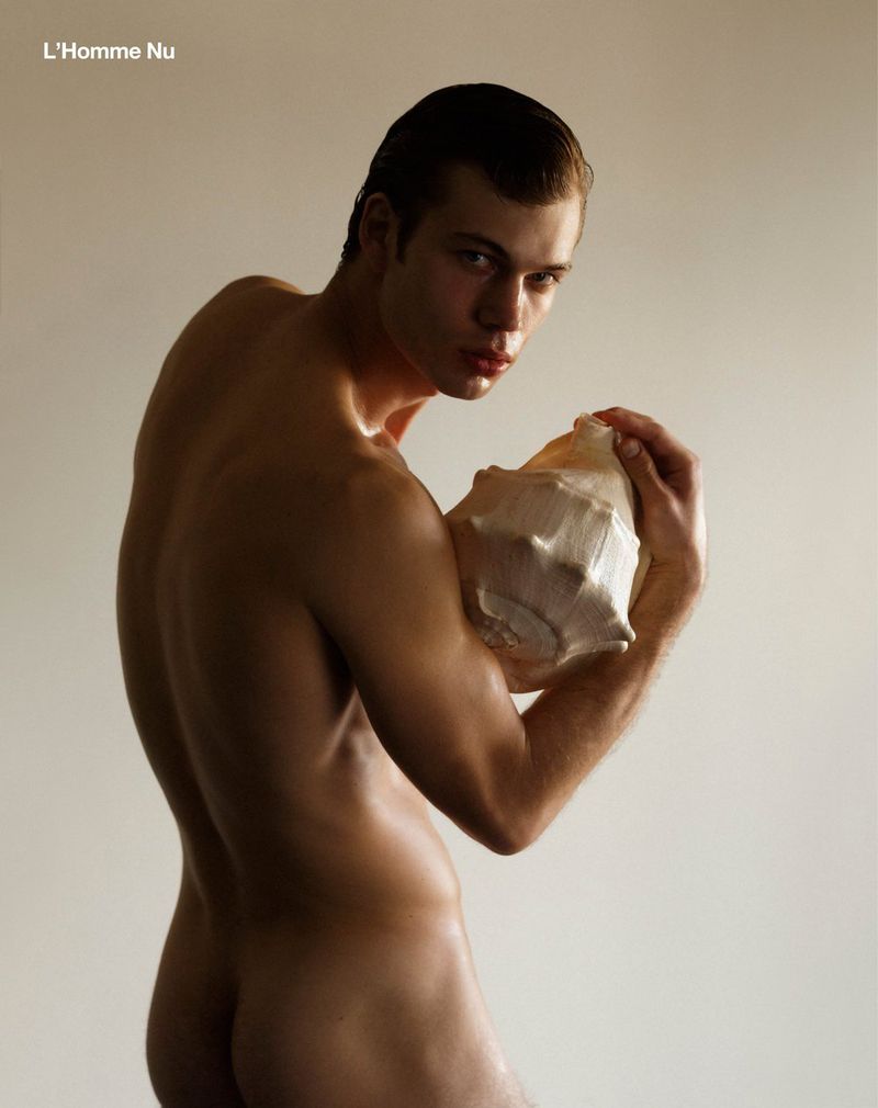 Nude male model poses in a studio with a large seashell in a portrait composition with soft neutral lighting and a clean aesthetic.