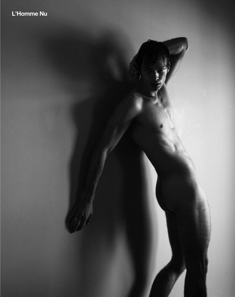 Monochrome portrait of a shirtless male model leaning against a wall in a studio with soft, diffused lighting. He is posing.