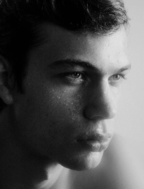 Monochrome portrait of a young man with short, dark hair and light eyes looking off to the side against a soft white background.