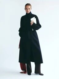 Model in dark coat holding books against white seamless background for ICICLE autumn winter 2026 campaign.