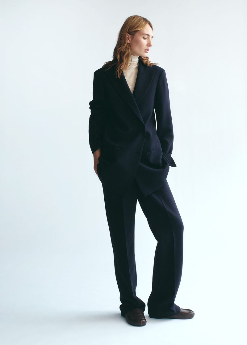 Model in a dark navy trouser suit and light turtleneck sweater standing against a clean white background.