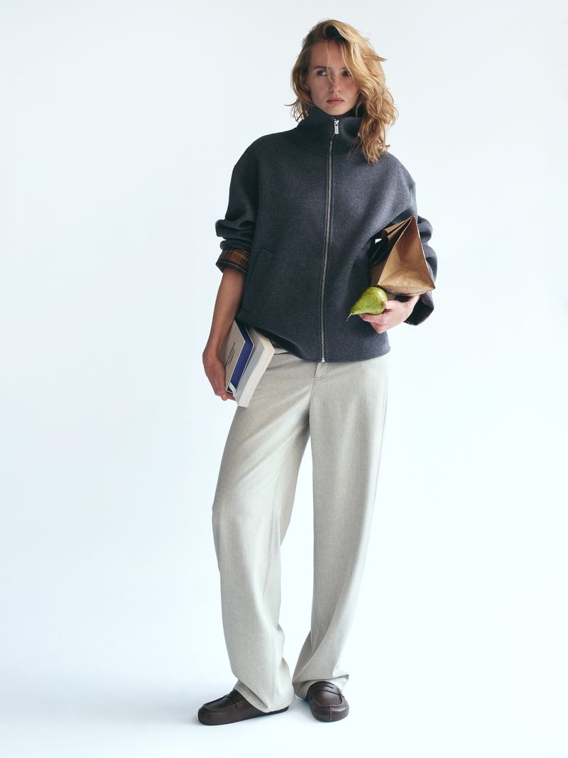 Model in a dark grey zip-up jacket and light grey trousers holding a book and a paper bag against a white seamless background