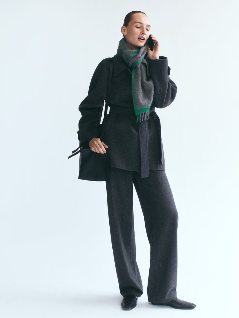 Model in dark wool coat and trousers wearing a green and grey scarf while holding a phone against a plain white background