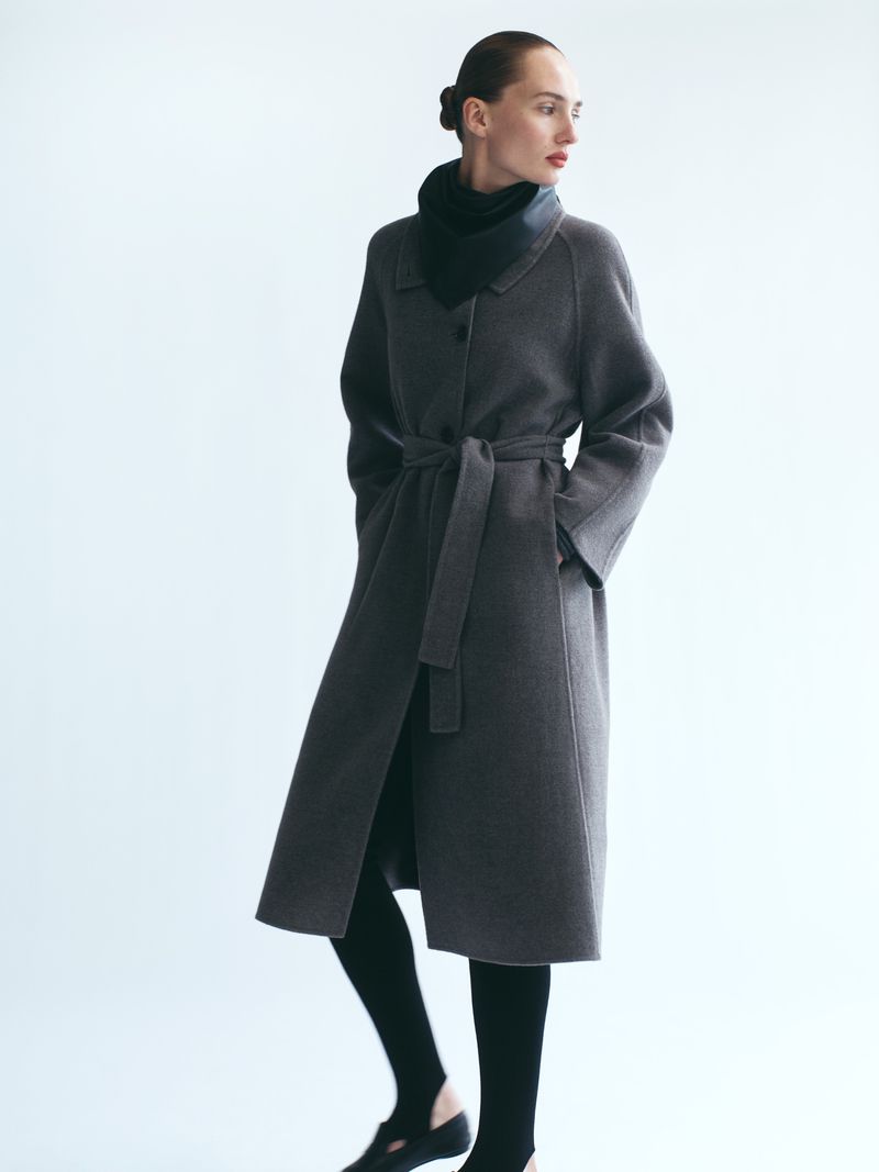 Model wearing a long grey belted wool coat and black accessories against a clean white background.