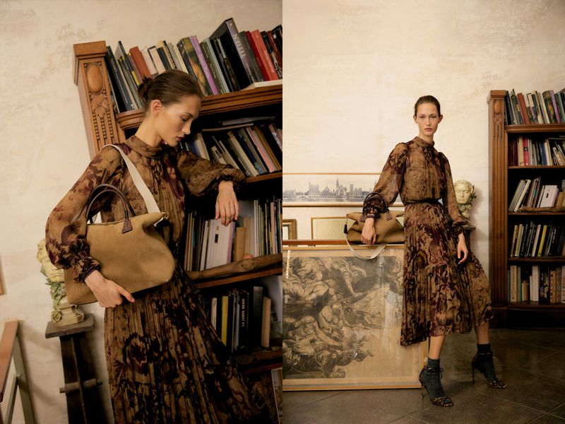 Model in brown patterned dress leaning against a wooden bookshelf holding a tan handbag in a library setting.