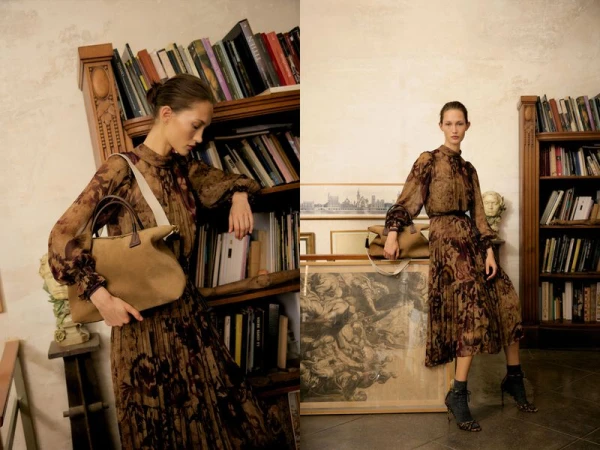 Model in brown patterned dress leaning against a wooden bookshelf holding a tan handbag in a library setting.